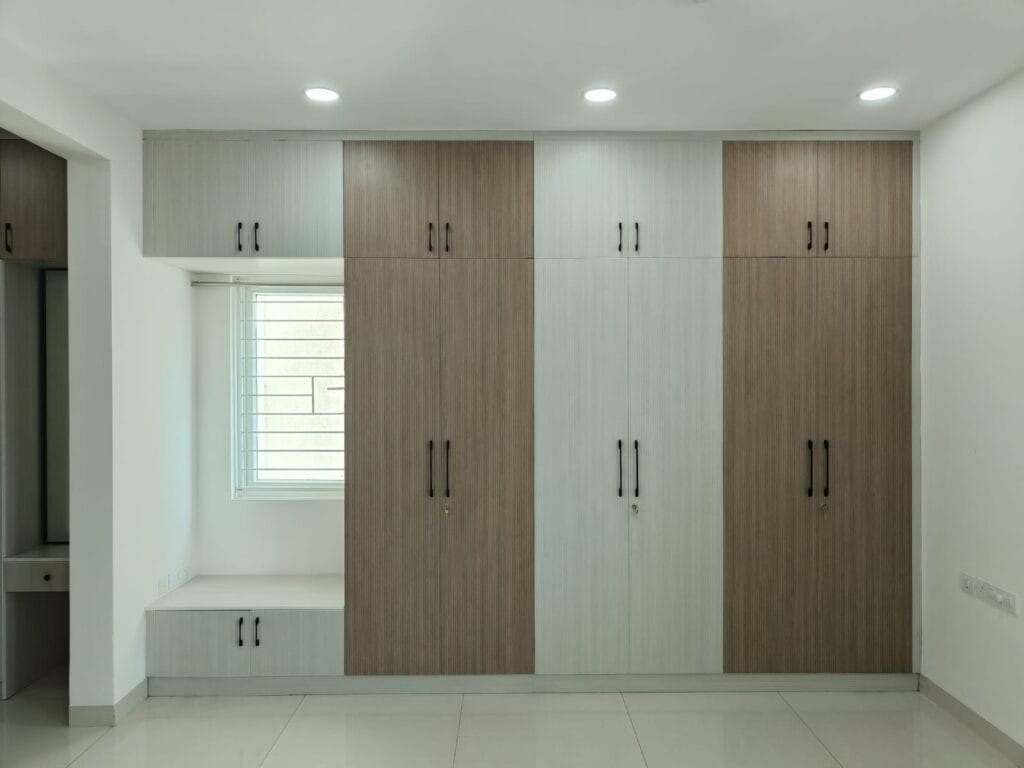 Four-door hinged wardrobe in brown wood finish with window seating by SB Interior Solutions Hyderabad